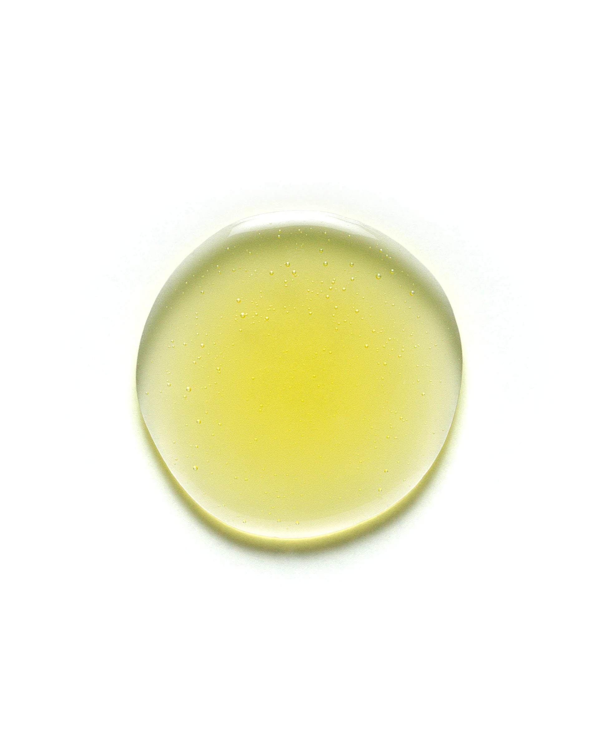 Close-up of a golden yellow droplet of Yerba Santa Micellar Cleansing Oil on a white background. The oil appears translucent with tiny suspended bubbles, showcasing its lightweight, hydrating texture. Skincare product image optimized for facial cleansing and makeup removal.